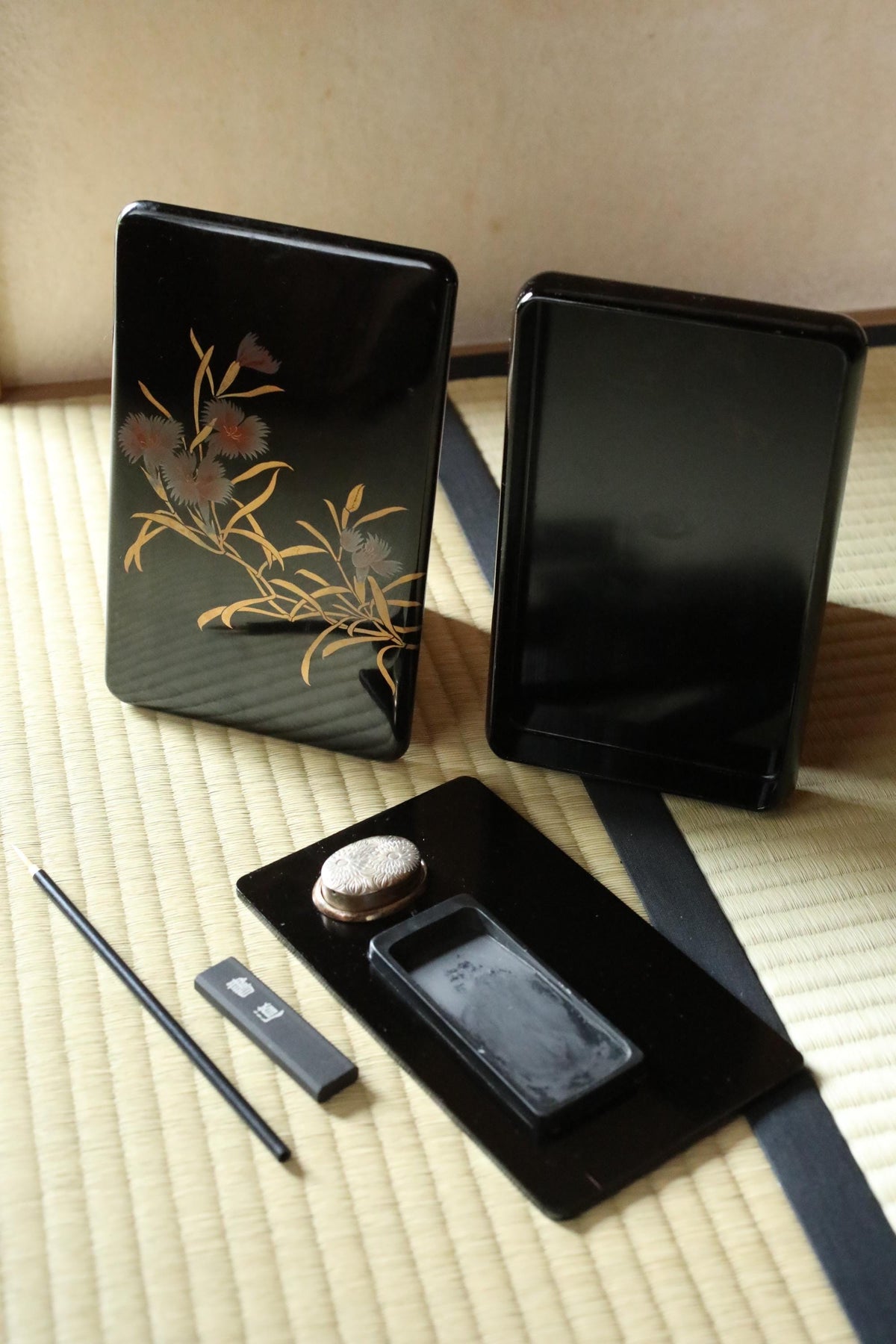 a pair of black lacquer boxes with floral designs, along with some other items on a woven mat.