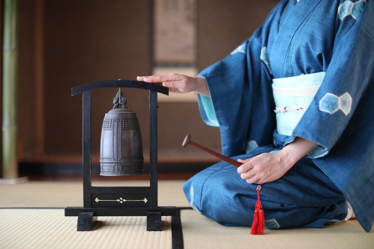 A person in traditional Japanese attire is kneeling and playing a large bell on a wooden stand, with a tassel hanging from the bell.