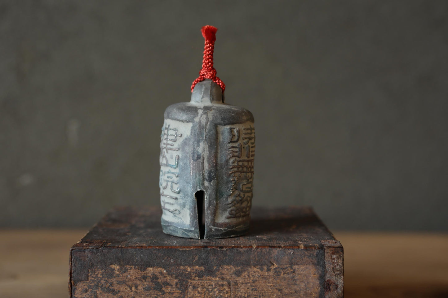A gray ceramic bell with a red string tied around it, sitting on a wooden surface against a gray background.