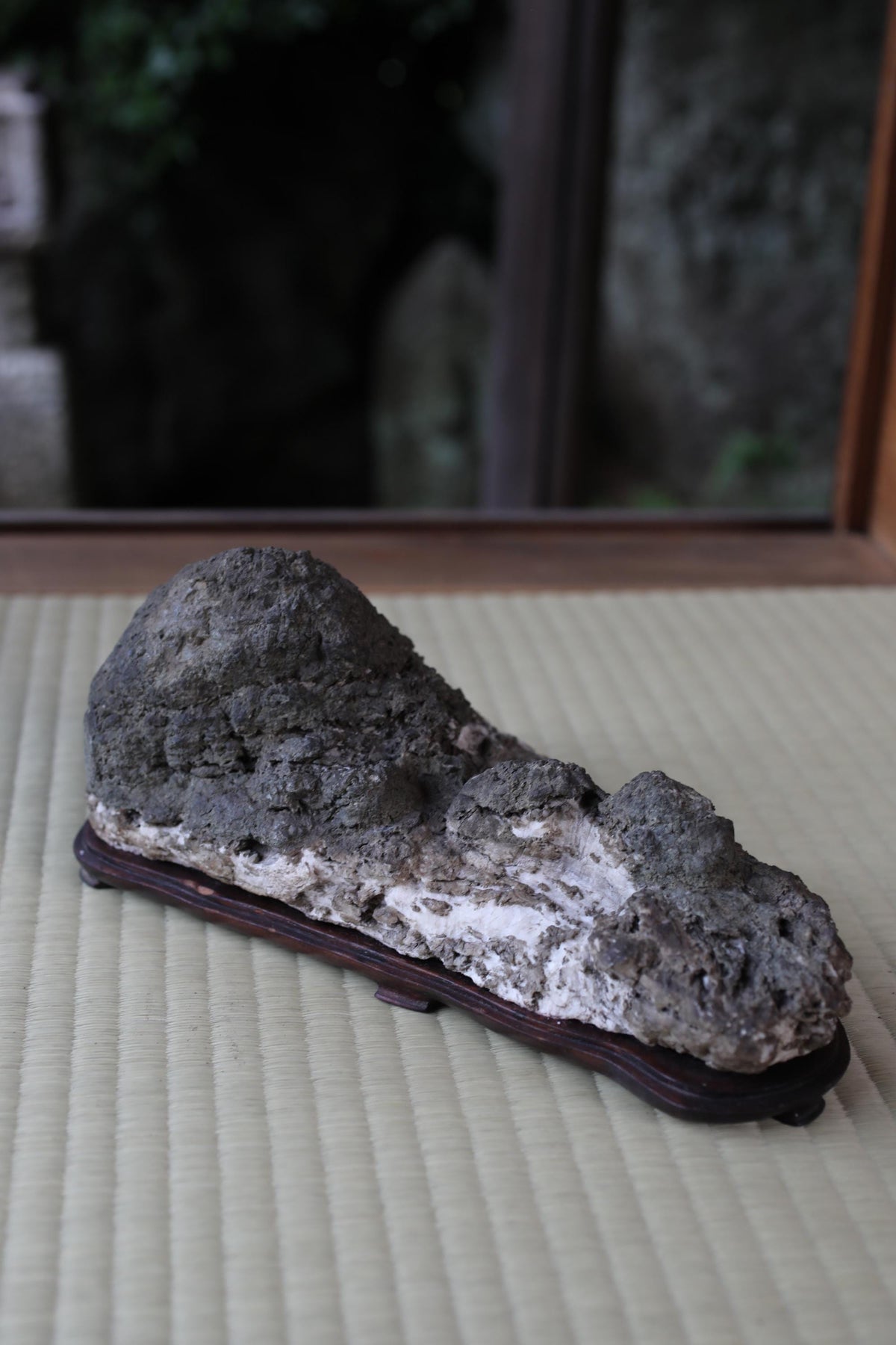 A large, irregularly shaped rock with a rough, textured surface sits on a wooden base, placed on a woven mat.