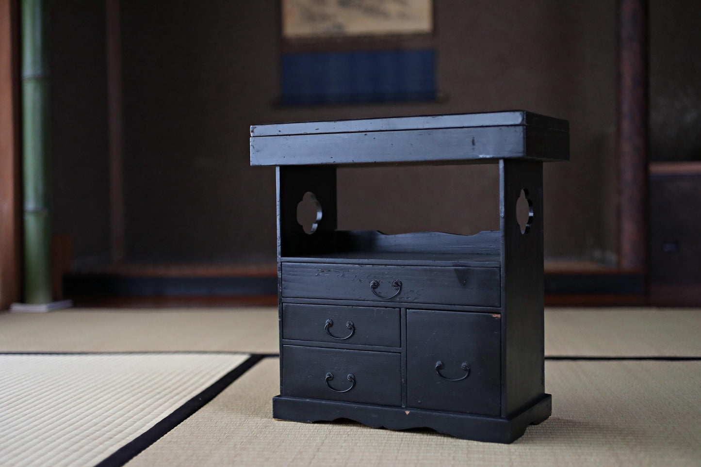 A small black wooden cabinet with three drawers and a shelf is placed on a tatami mat in a room with a wooden wall and a window.