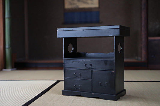 A small black wooden cabinet with three drawers and a shelf is placed on a tatami mat in a room with a wooden wall and a window.