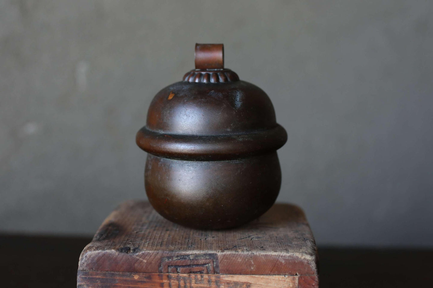 A dark brown, round metal container with a lid sits on a wooden surface against a gray background.