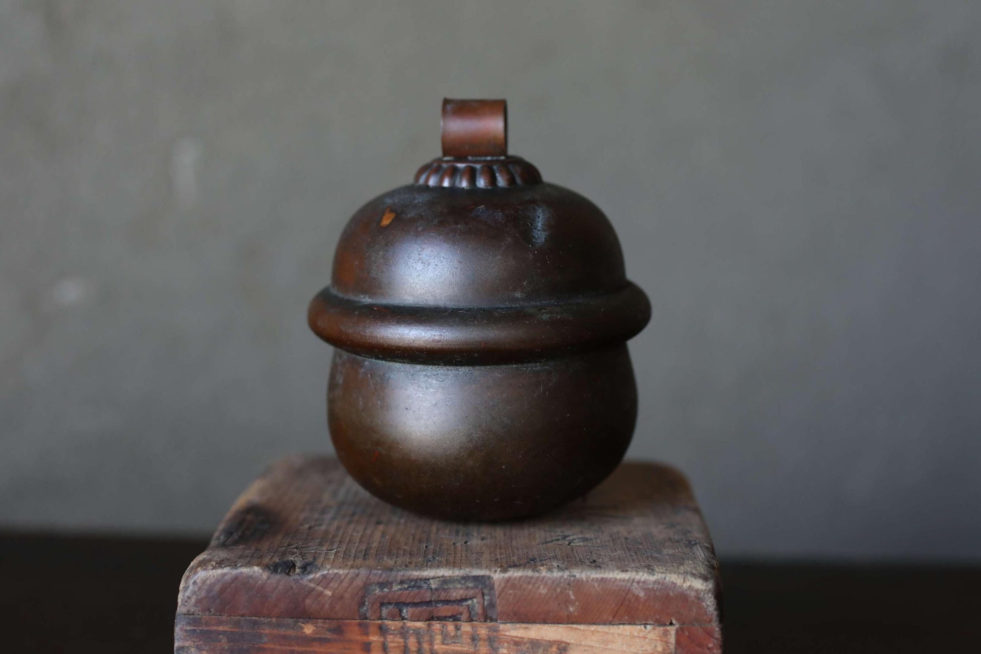 A dark brown, round metal container with a lid sits on a wooden surface against a gray background.