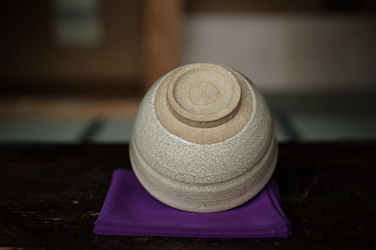 A white ceramic bowl with a round base and a small circular design sits on a purple cloth on a dark surface.
