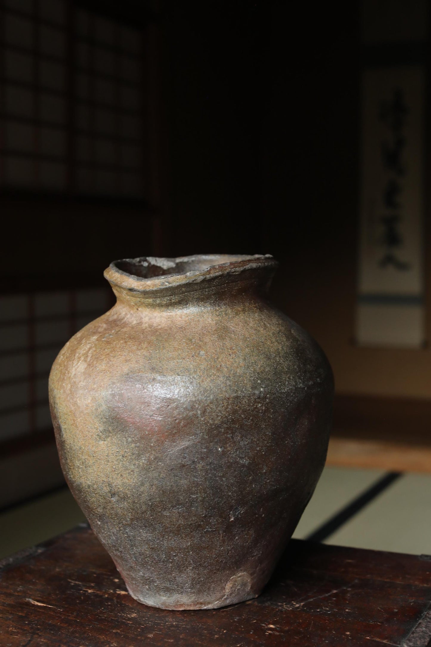 An old, weathered ceramic vase sits on a wooden surface, with a blurred background that includes a traditional Japanese-style wall.