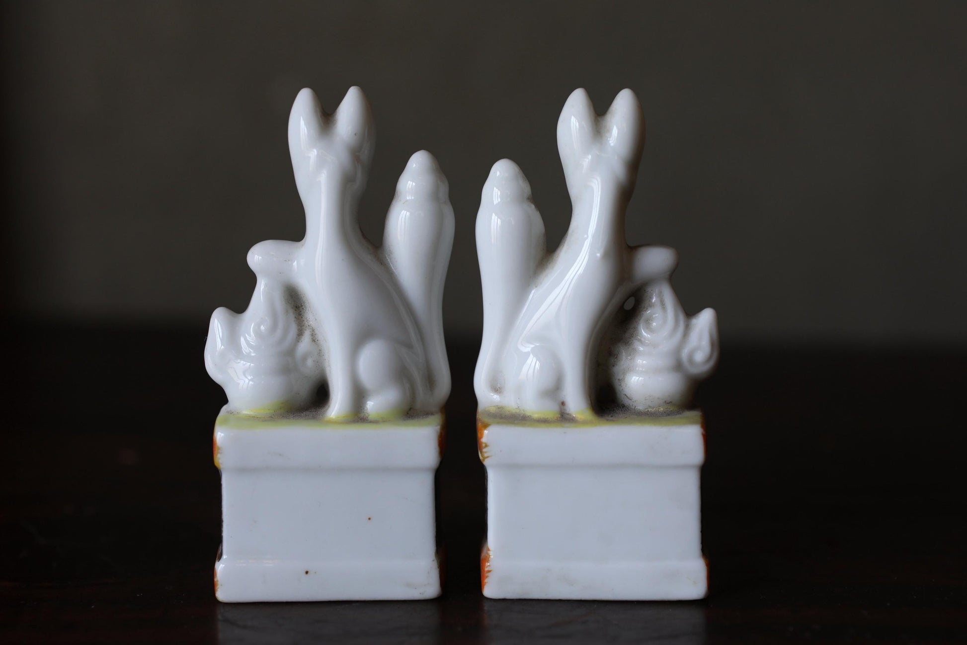 Two white ceramic figurines of cats sitting on pedestals against a dark background.