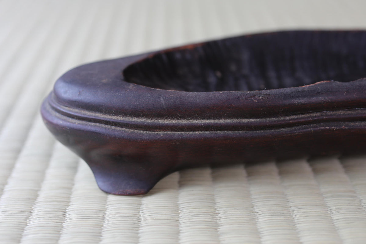 A dark brown, ornate vase or container rests on a woven surface.