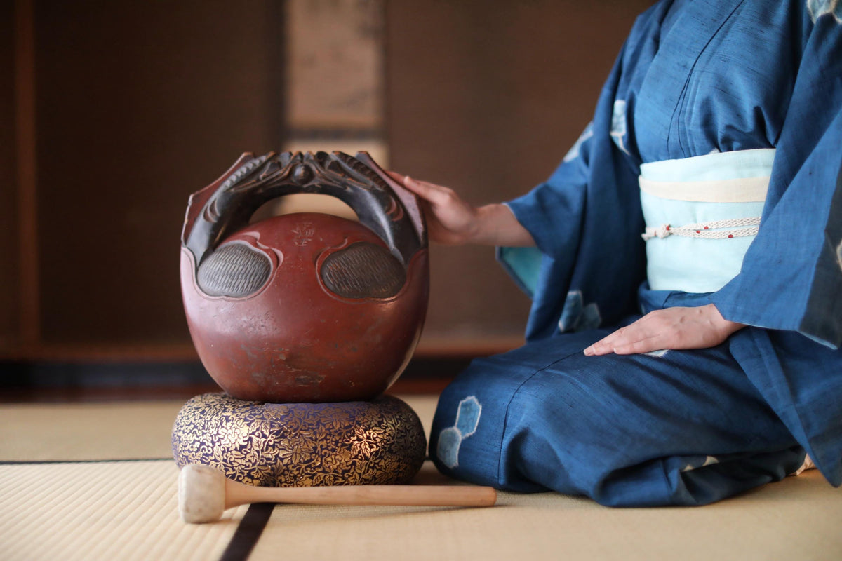 A person in a blue kimono is seated on the floor, touching a red and black ceramic object with a wooden stick.