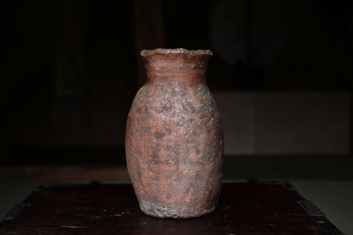 An old, weathered clay vase sits on a dark wooden surface, its reddish-brown color and rough texture suggesting it has been used for an extended period.