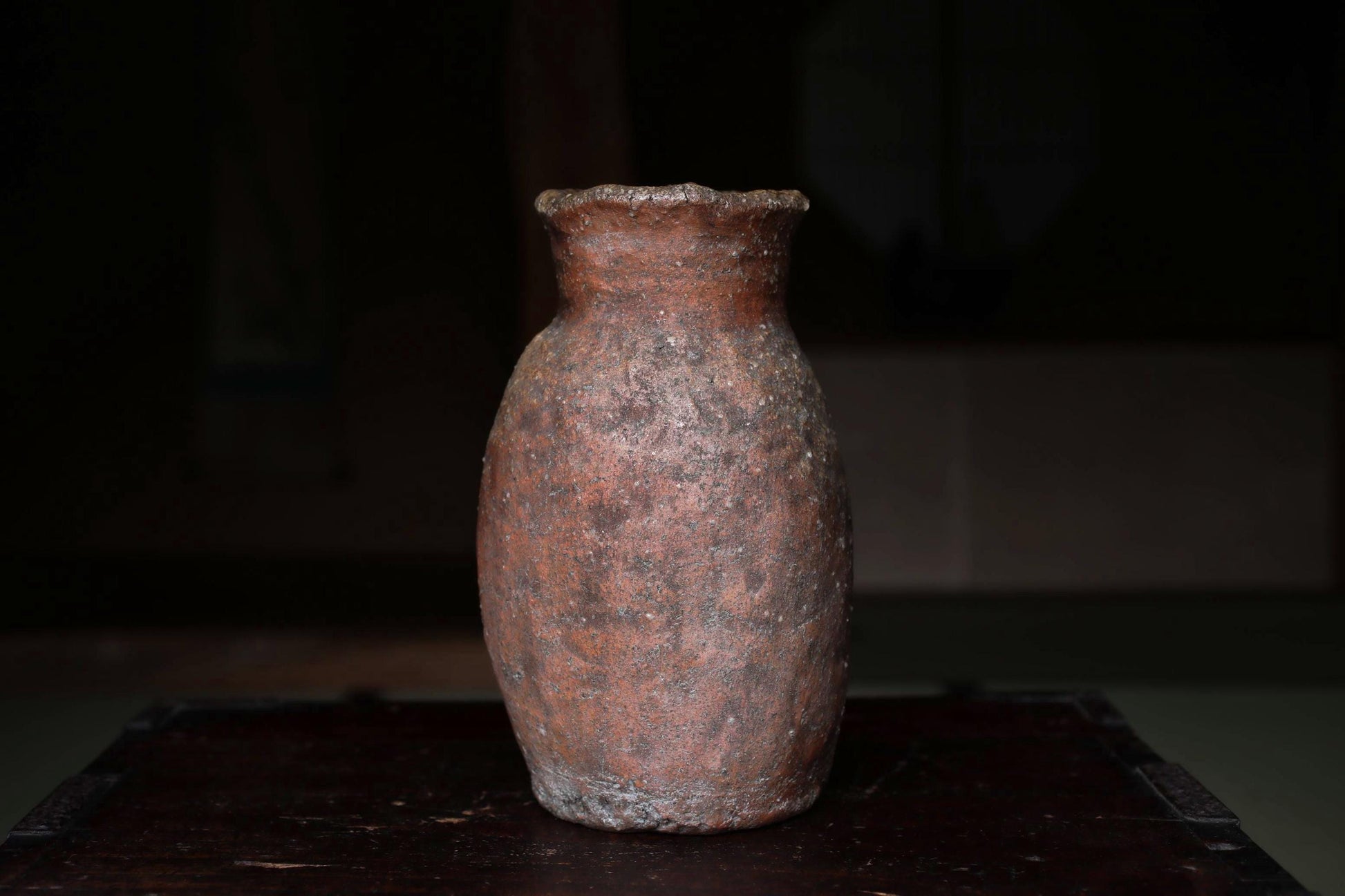 An old, weathered clay vase sits on a dark wooden surface, its reddish-brown color and rough texture suggesting it has been used for an extended period.
