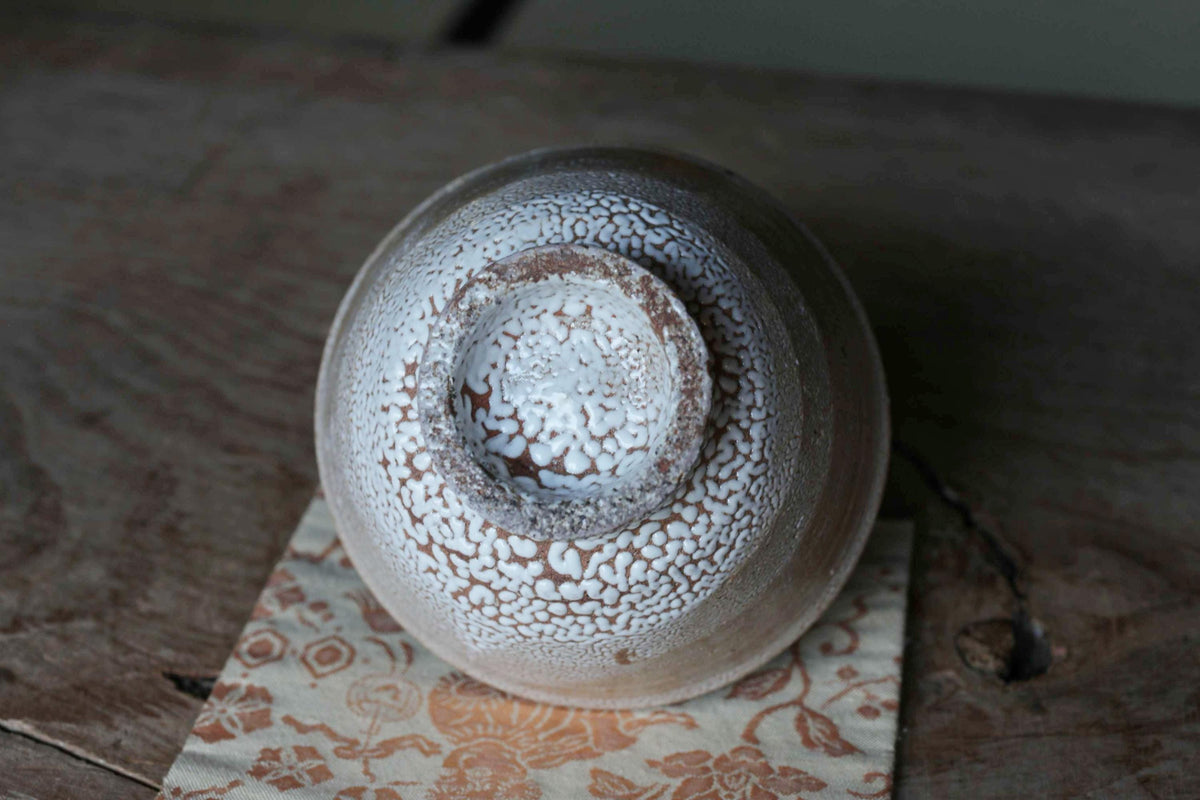 A ceramic bowl with a white, speckled pattern sits on a wooden surface, with a floral patterned cloth underneath.