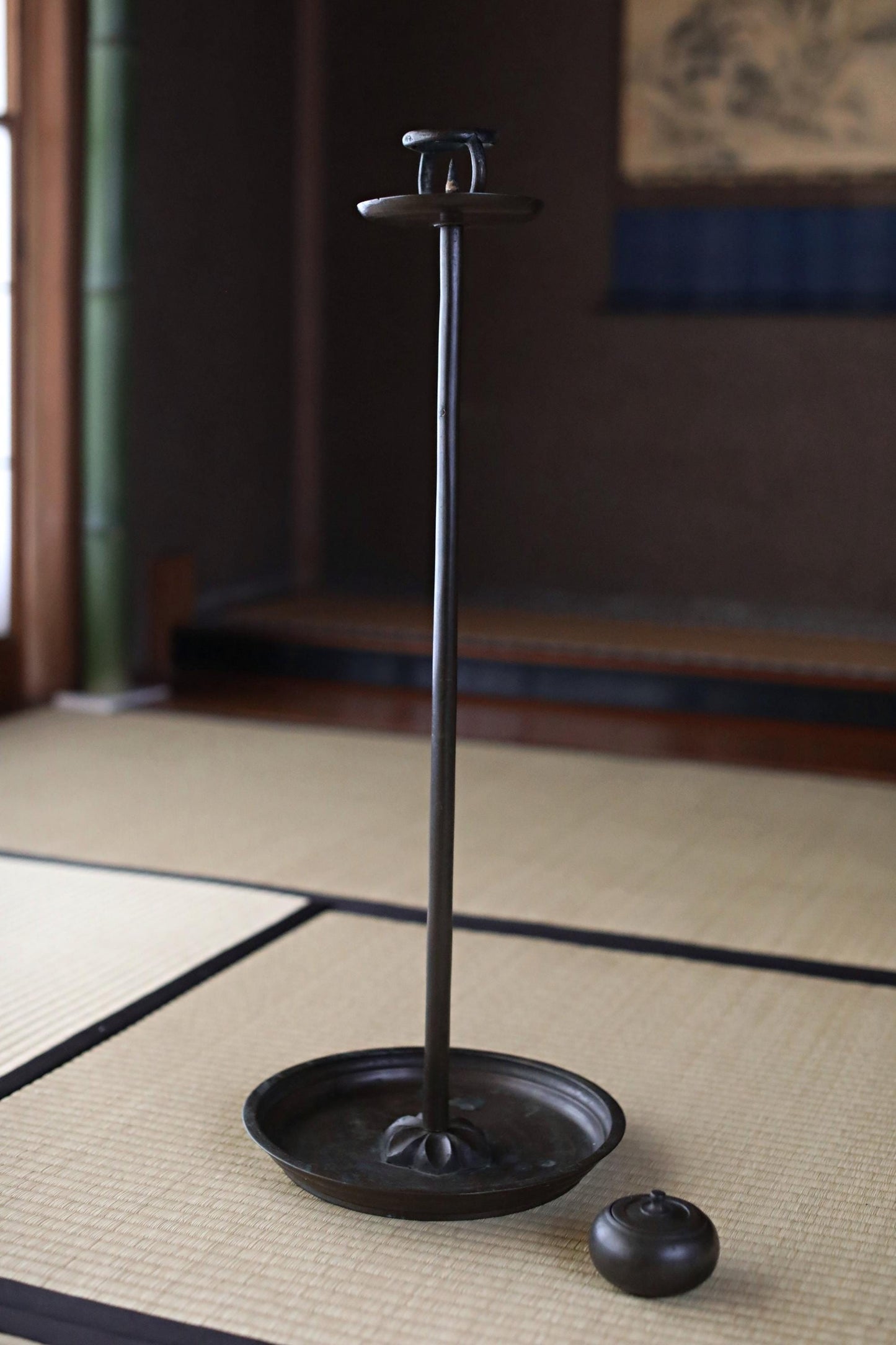 A black metal candle holder with a candle sits on a woven mat in a dimly lit room.