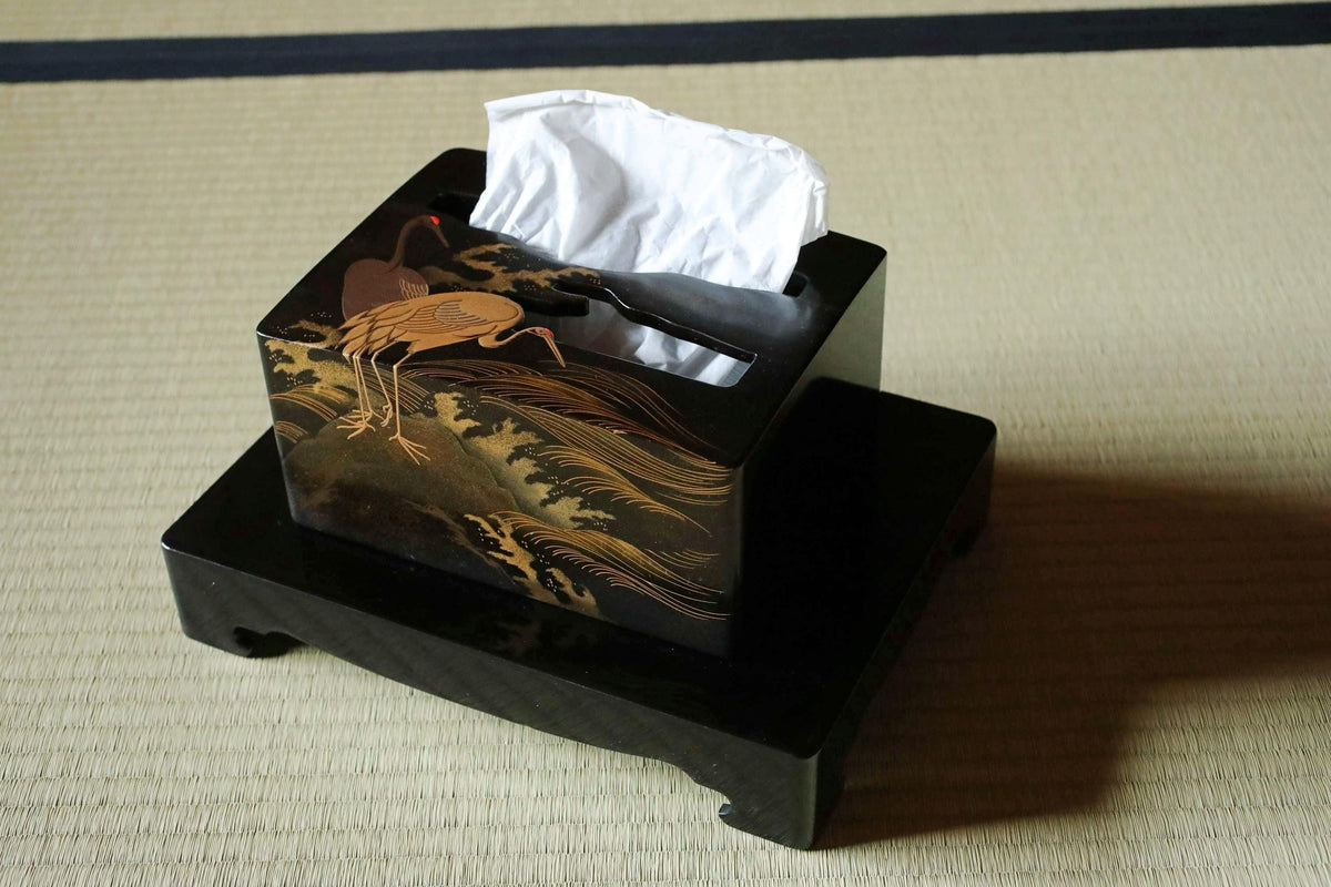 A black tissue box with a gold and black design depicting a crane on a bamboo mat, placed on a black stand.