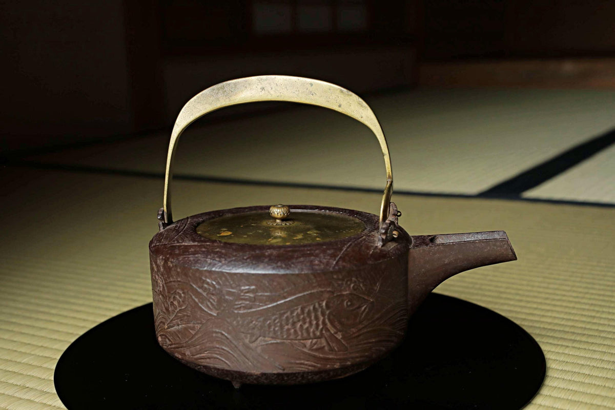 A brown, ornately decorated teapot with a gold handle and spout sits on a black surface, likely a table or mat, in a dimly lit room.