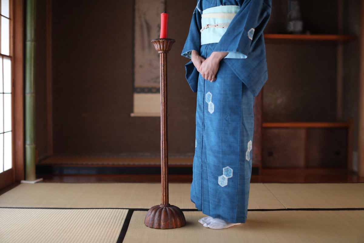 A person wearing a traditional Japanese kimono stands in a room with a tatami mat, a wooden candlestick, and a window.