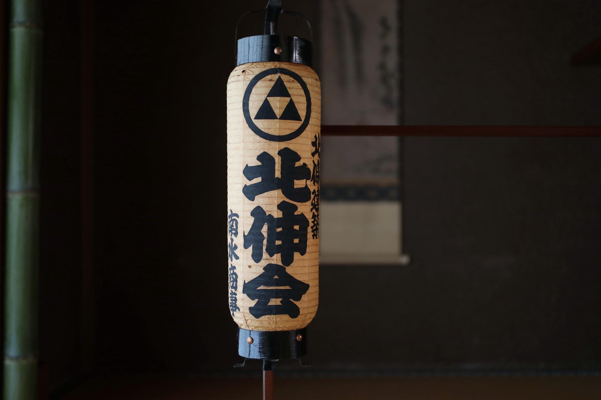 A traditional Japanese paper lantern with black text and a triangular symbol on it, hanging in a dark setting.