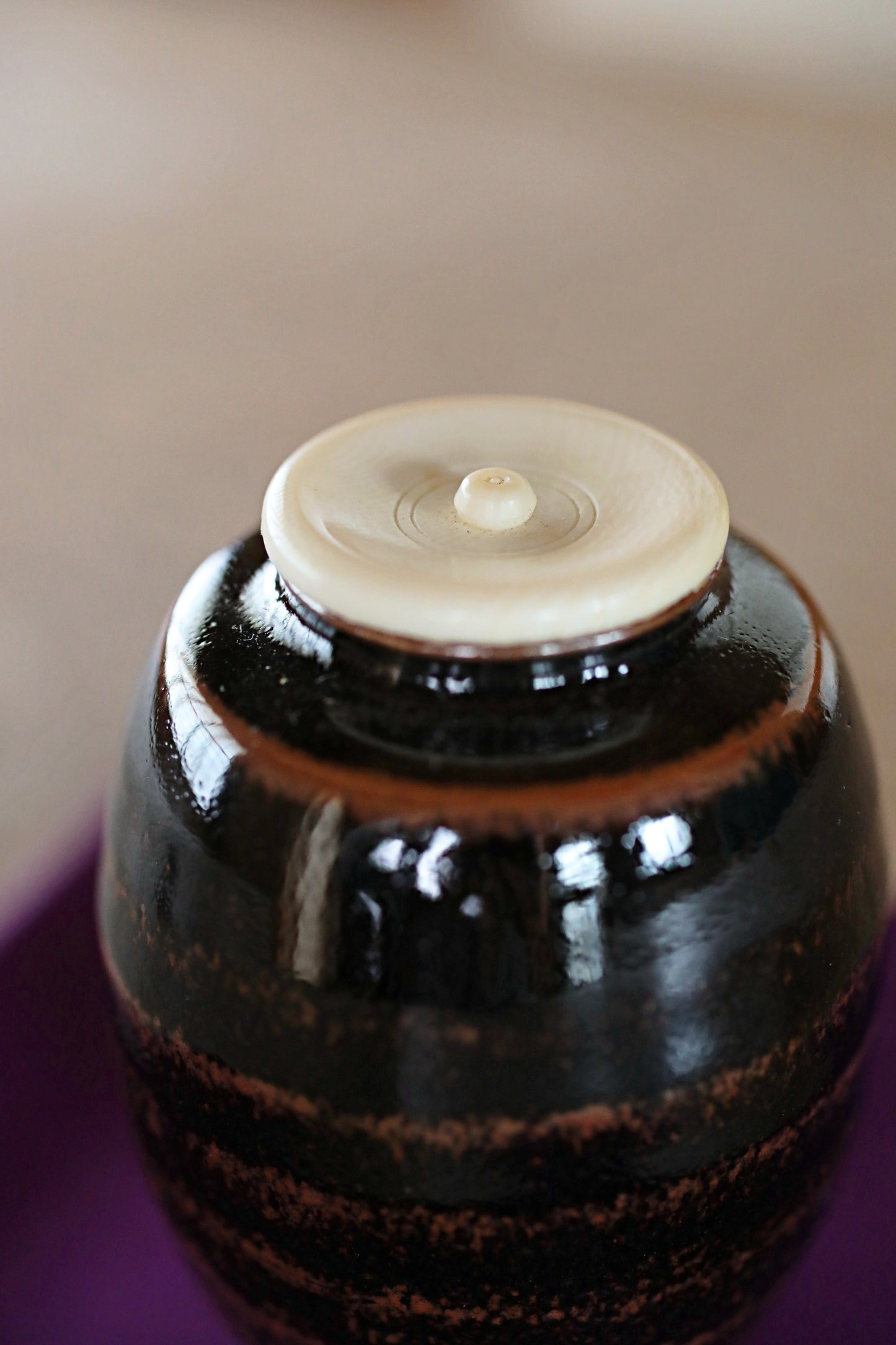A close-up image of a dark brown ceramic vase with a white lid, placed on a purple surface.