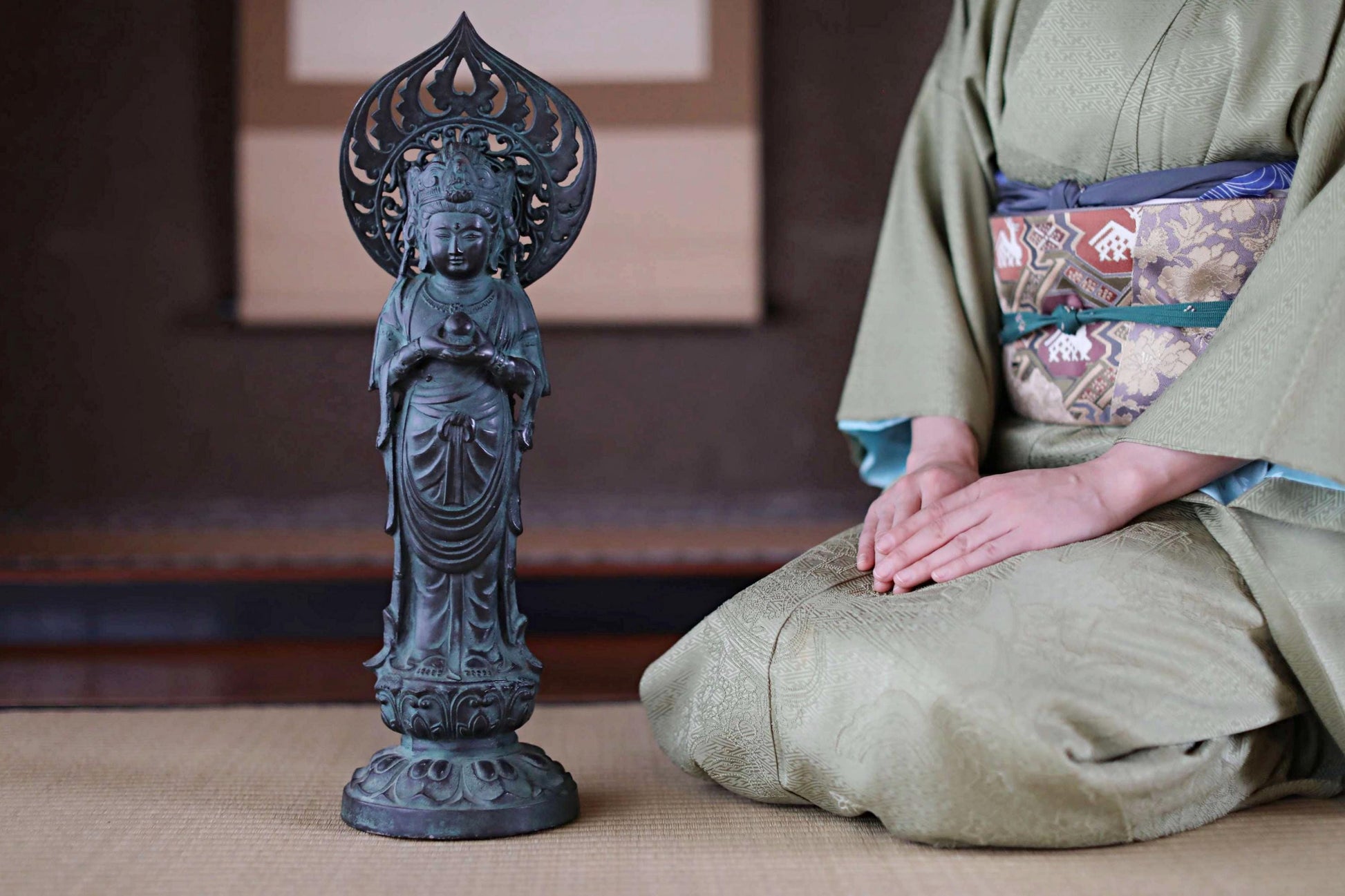 A person in traditional Japanese attire, including a kimono, is kneeling next to a small statue of a deity, likely a Buddha, in a room with a tatami mat.