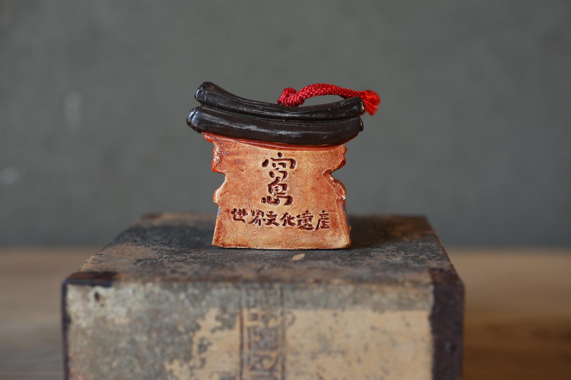 A ceramic figurine with a red string and a black object on top, sitting on a wooden surface against a gray background.