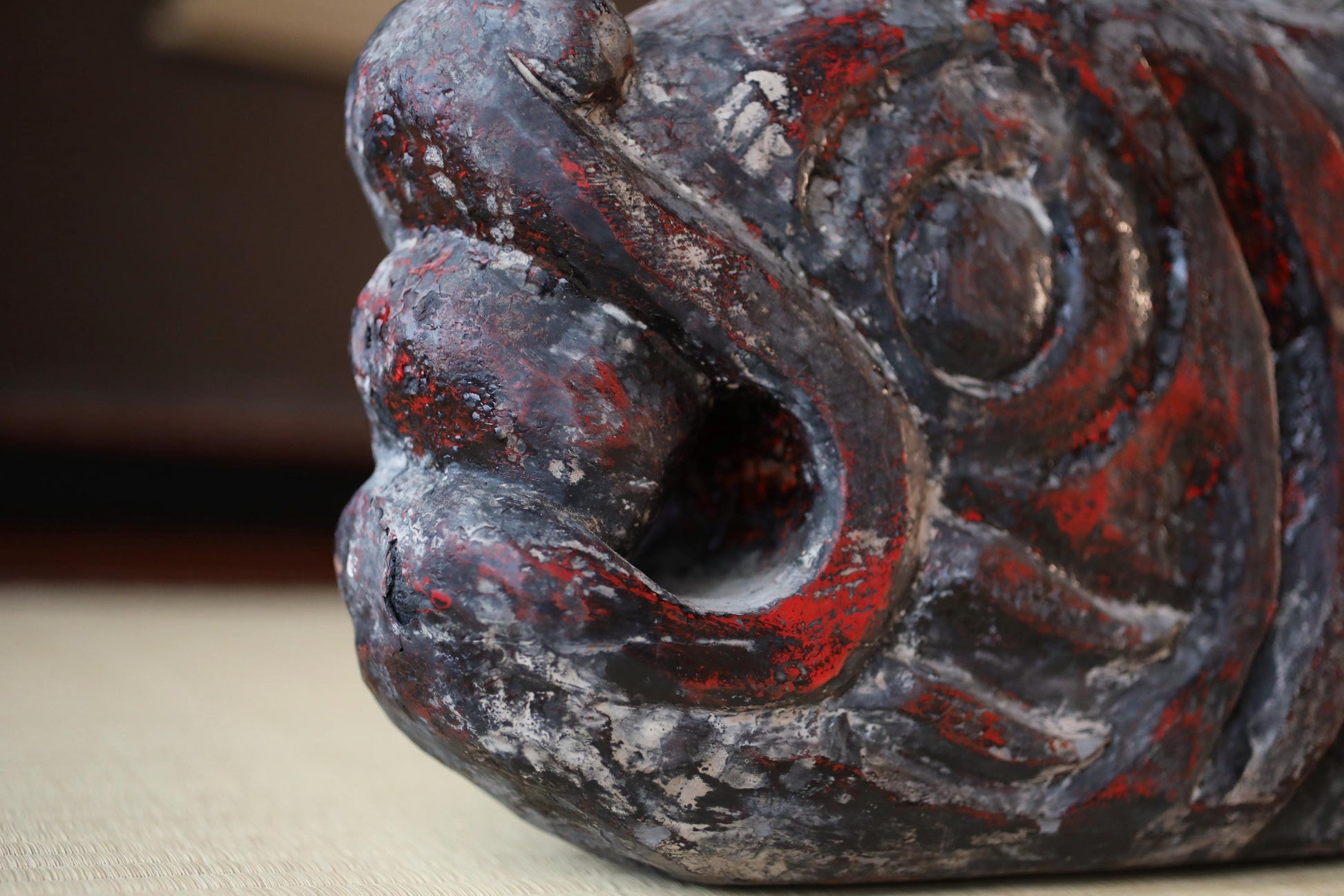 A close-up view of a weathered, dark-colored sculpture with a red and black patterned texture, sitting on a surface.