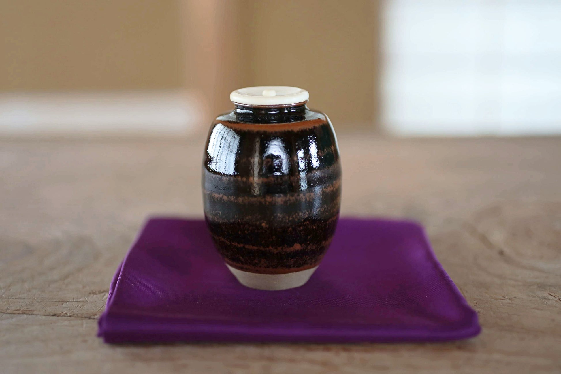 A small, round ceramic jar with a white lid sits on a purple cloth on a wooden surface.