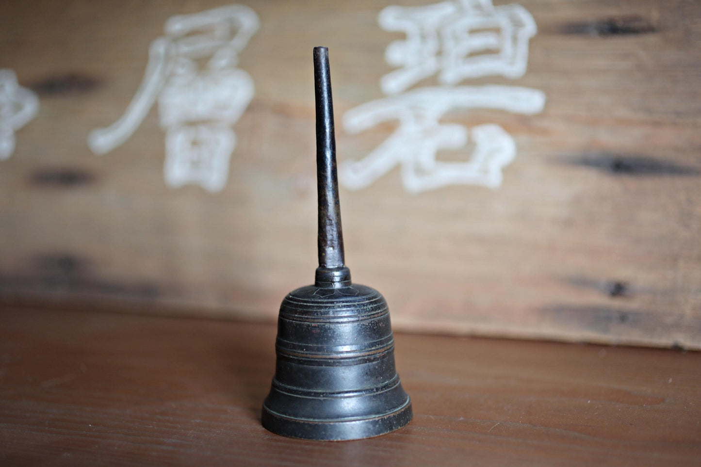 A black metal bell is placed on a wooden surface, with a wooden wall in the background featuring white characters.