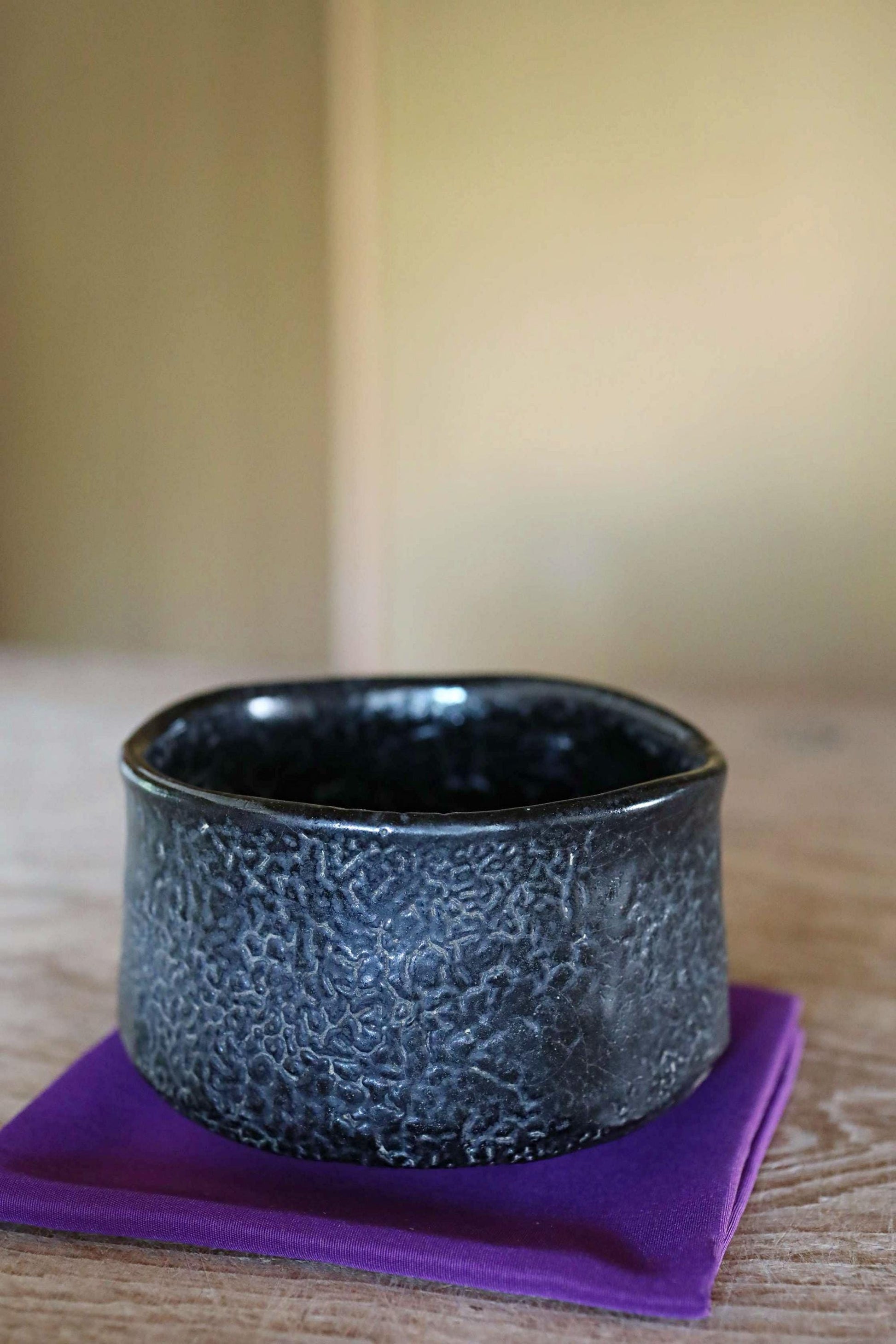 A black ceramic bowl with a textured surface sits on a purple cloth on a wooden surface.