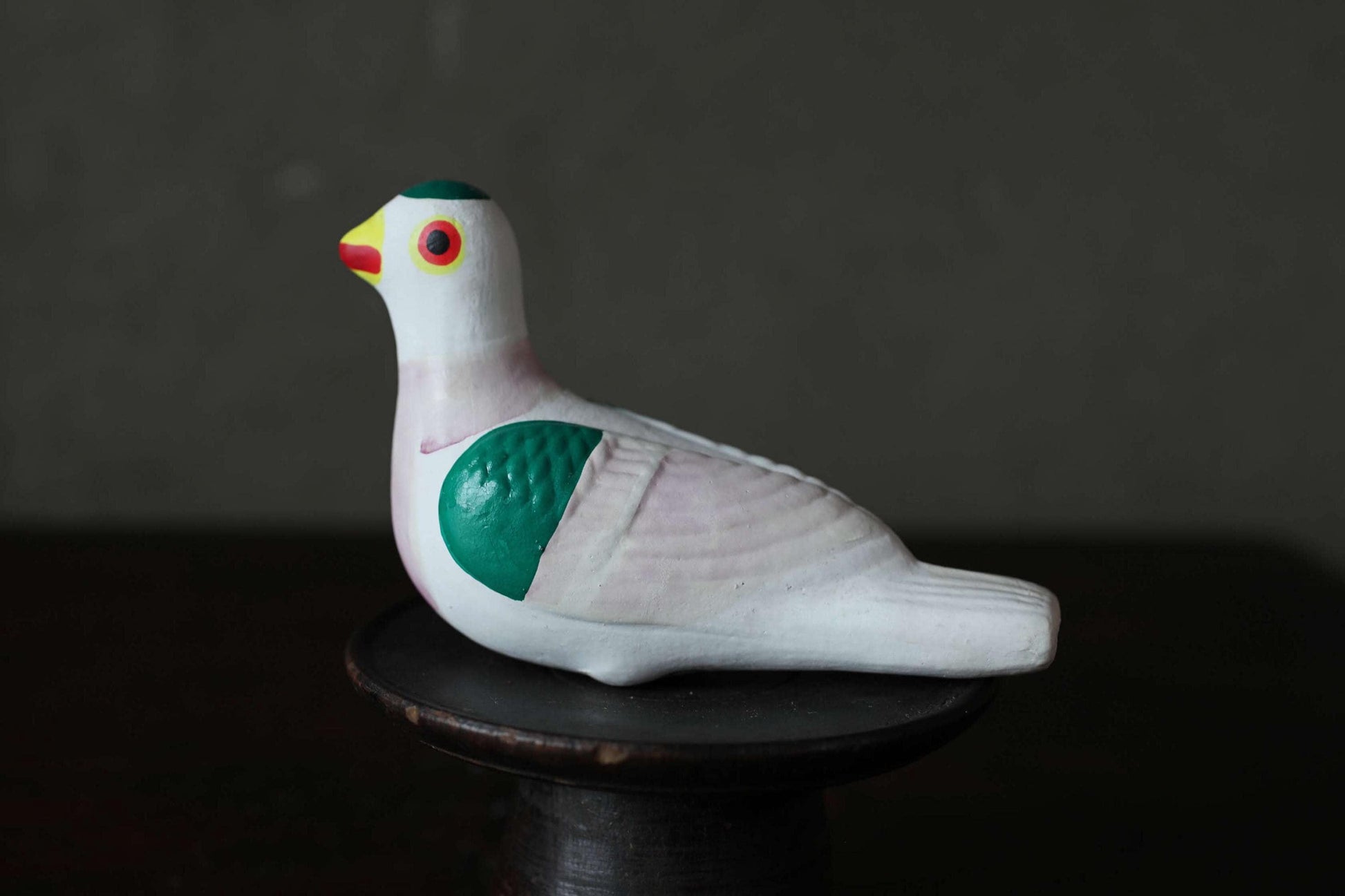 A ceramic bird figurine with a white body, green wings, and a red beak is sitting on a black stand against a dark background.