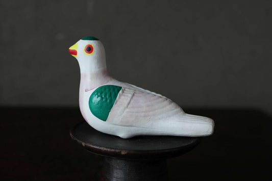 A ceramic bird figurine with a white body, green wings, and a red beak is sitting on a black stand against a dark background.