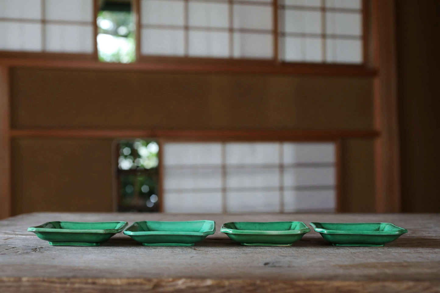Four green square bowls are arranged on a wooden table, with a blurred background that includes a window and a wooden wall.