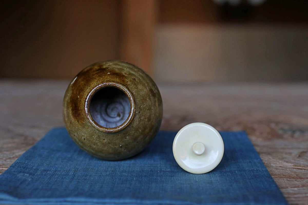 A brown ceramic jar with a spiral design sits on a blue cloth, accompanied by a white lid, on a wooden surface.