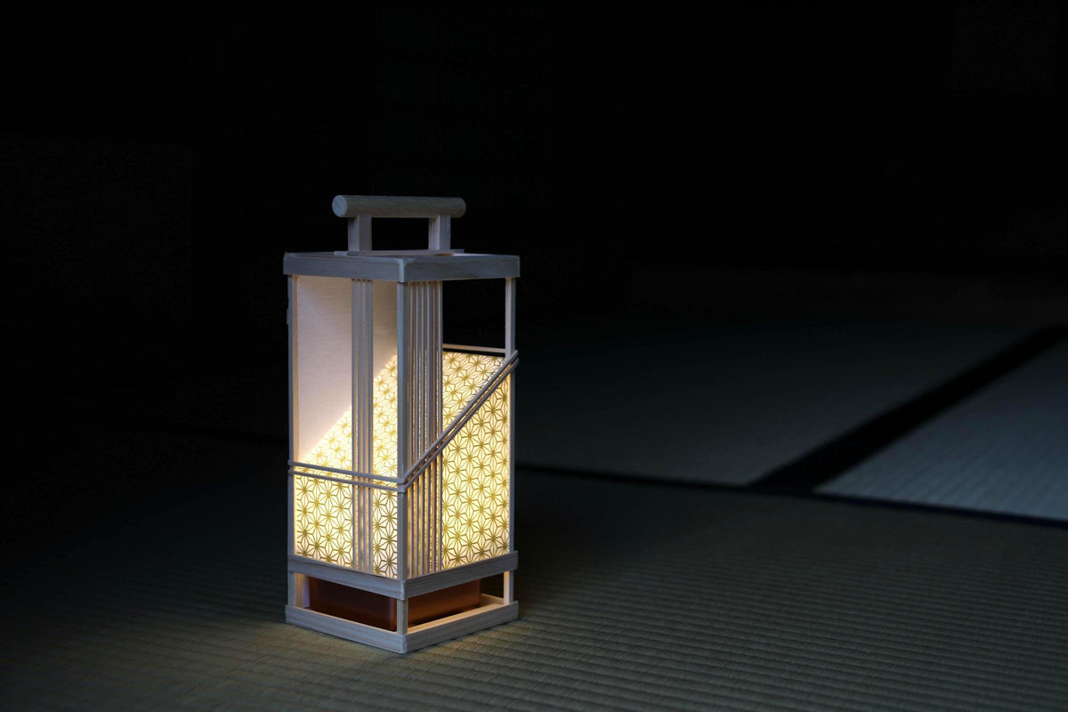 A white lantern with a handle and a patterned glass cover is illuminated in a dark room, casting a warm glow.
