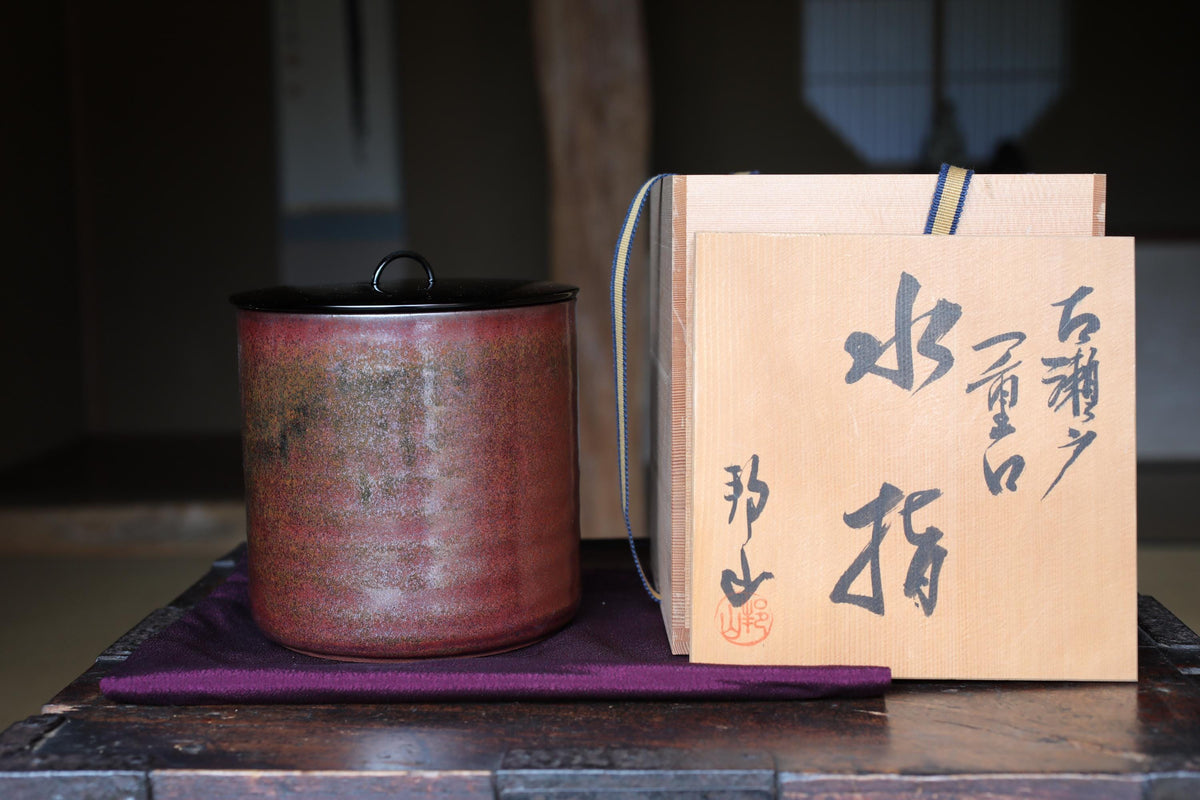 a red ceramic pot with a black lid, placed on a wooden surface next to a wooden box with Japanese characters on it.