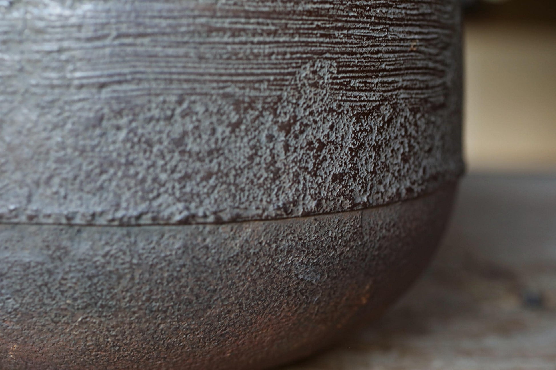A close-up view of a textured, weathered surface, possibly a piece of pottery or a metal object, with a rough, uneven texture and a dark, muted color palette.