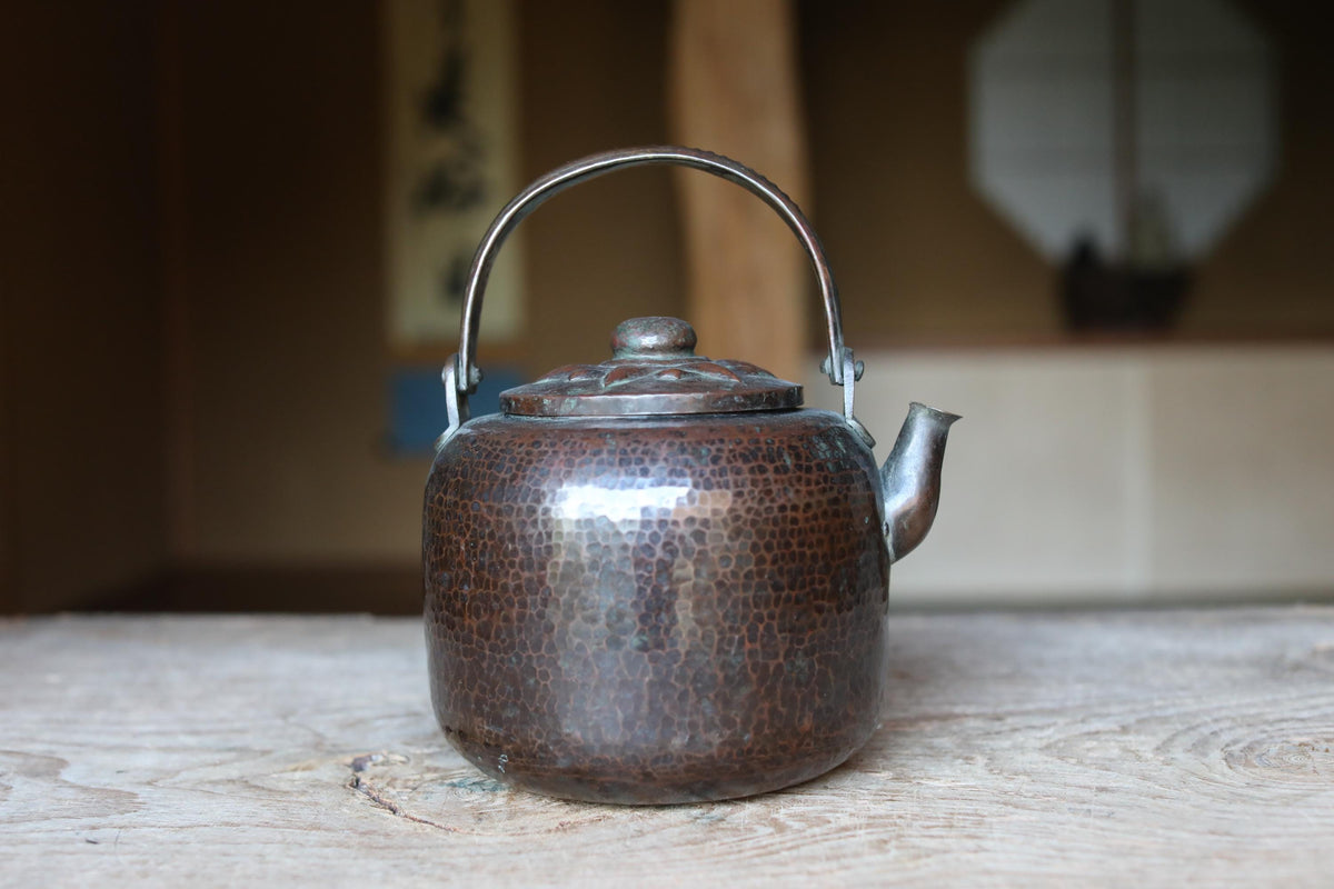 A shiny, metallic teapot with a handle and spout sits on a wooden surface, with a blurred background that includes a window and a plant.