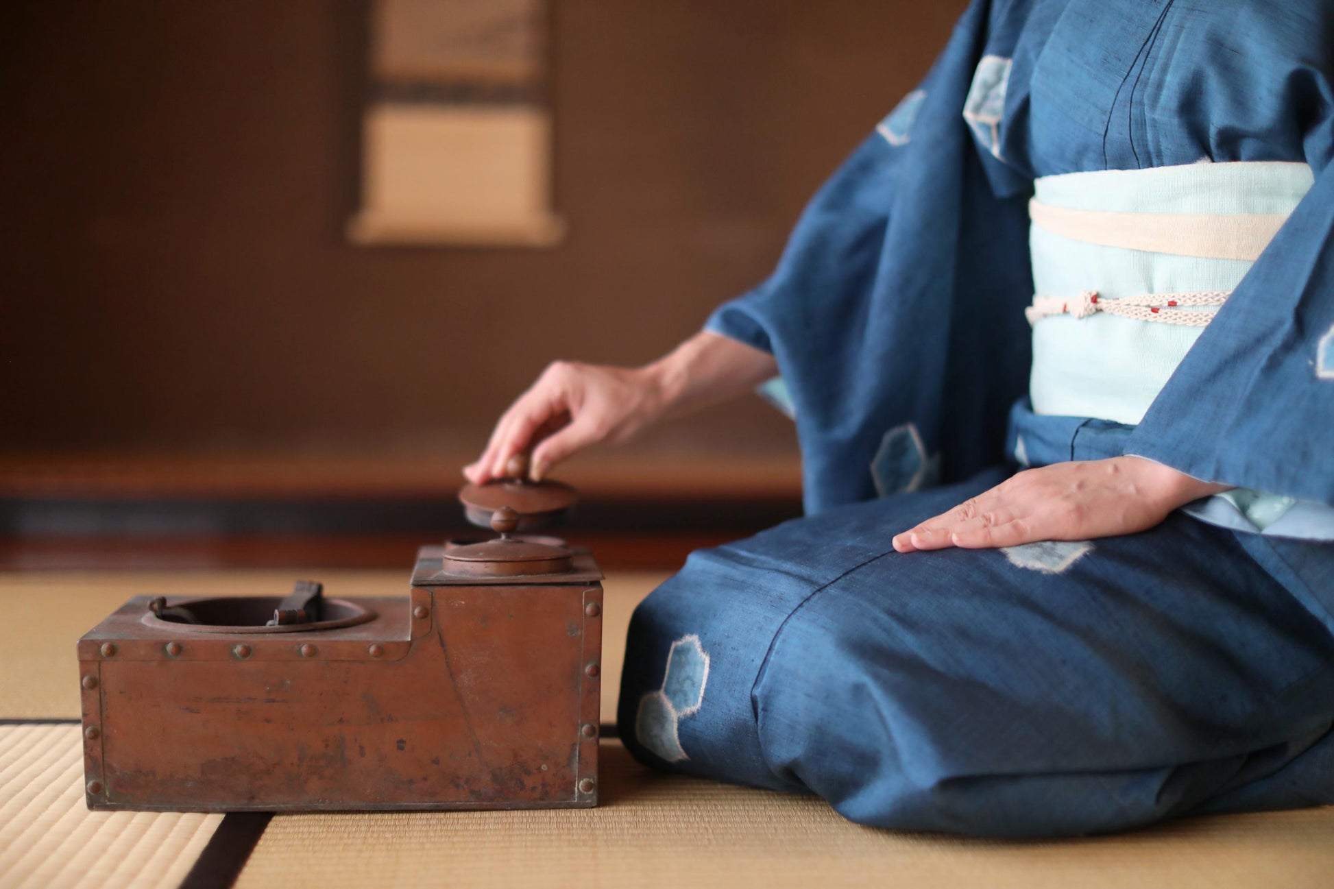 A person in a blue kimono is sitting on the floor, holding a small wooden object, possibly a tea pot or a small wooden box.