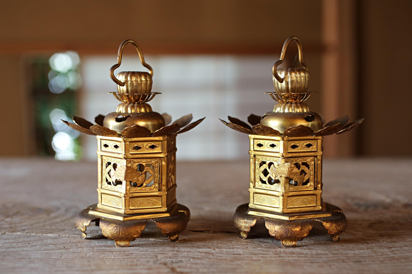 Two ornate brass lanterns with intricate designs sit on a wooden surface, their golden hues contrasting with the natural tones of the wood.