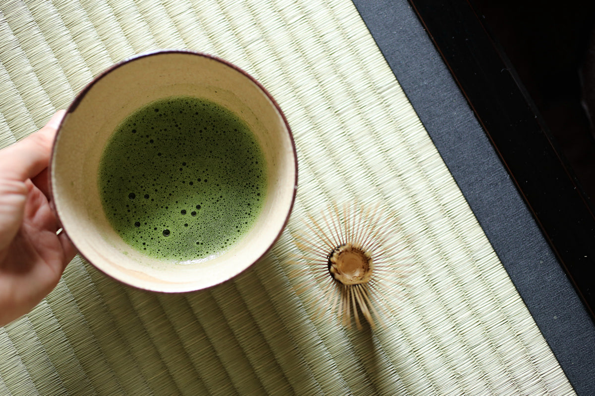 A hand holds a cup of green tea next to a wooden tea strainer on a woven mat.
