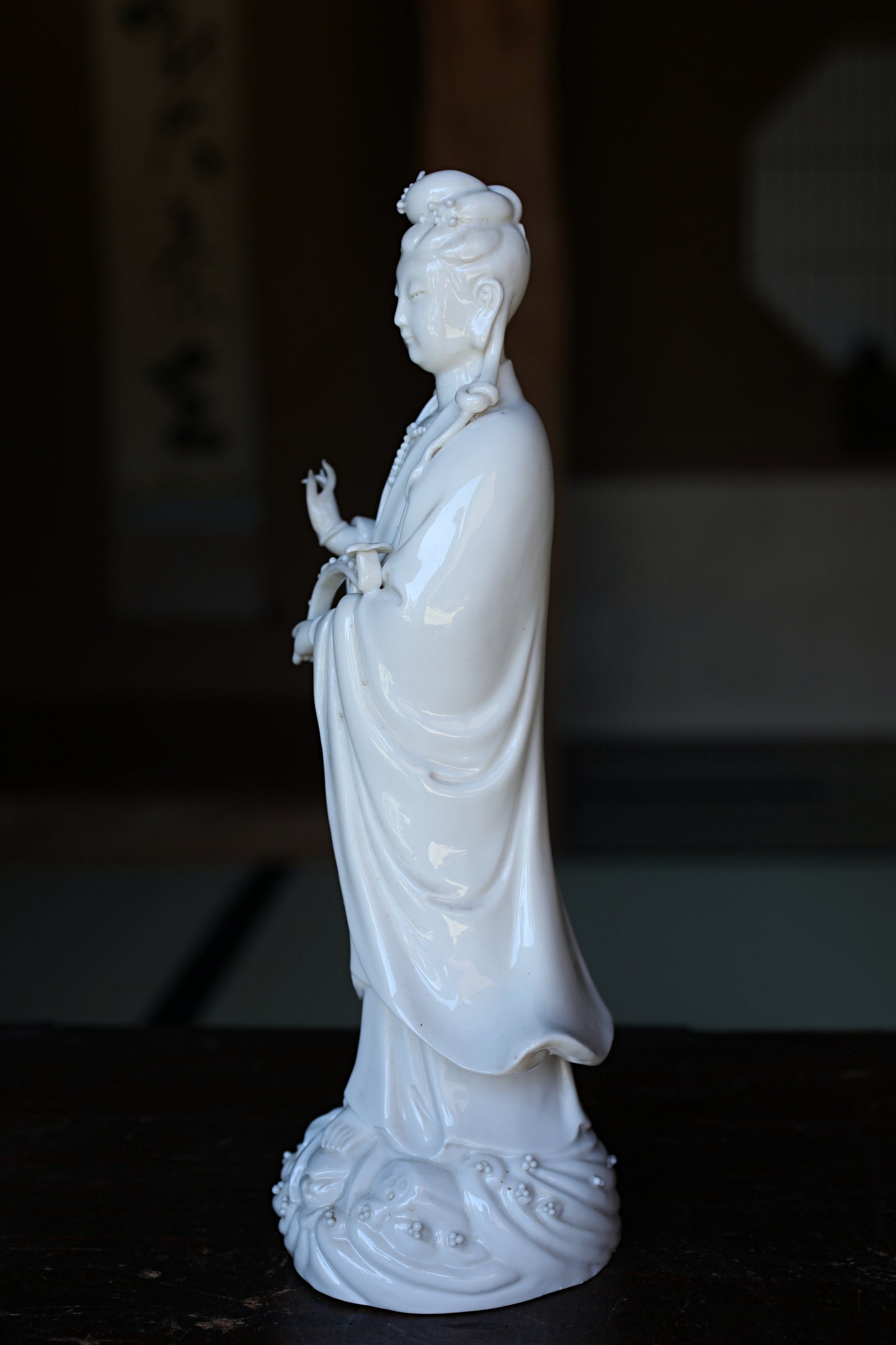 A white ceramic statue of a person standing on a base with intricate designs, placed on a dark surface.