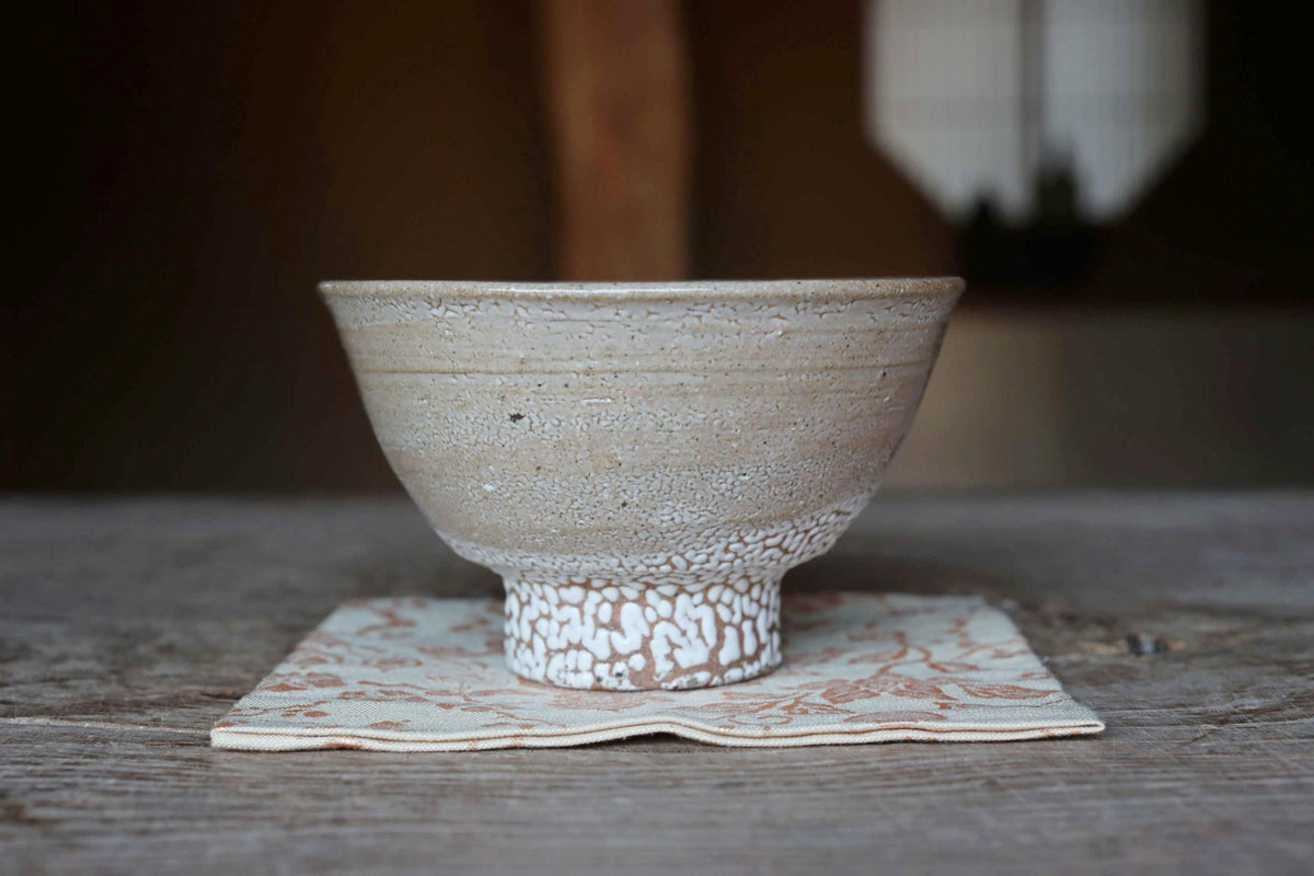 A white ceramic bowl with a speckled pattern sits on a white and brown floral napkin on a wooden table.