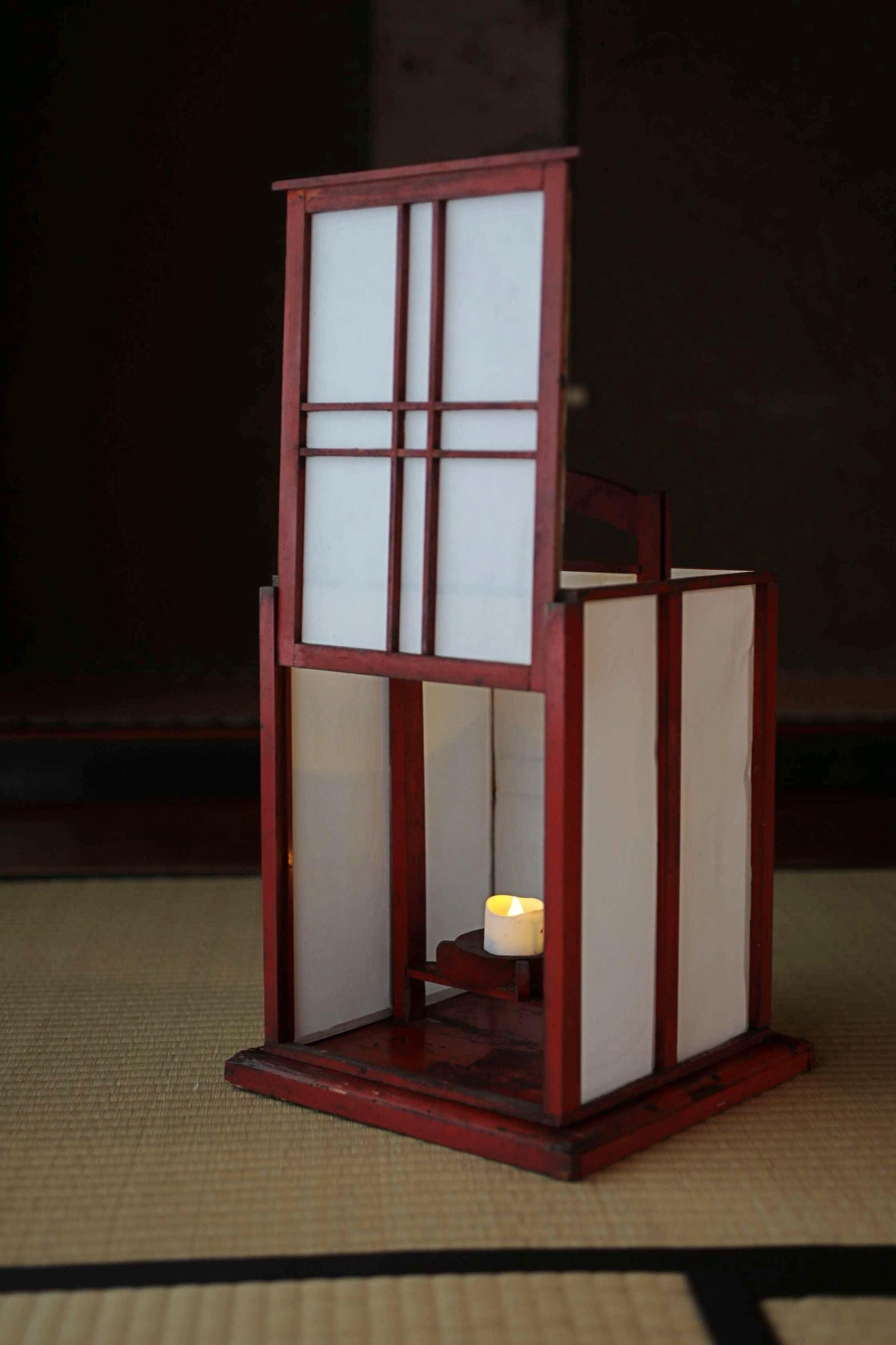A red wooden lantern with a white interior sits on a tatami mat, casting a warm glow.