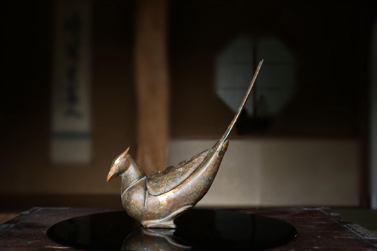 A bronze bird figurine is displayed on a reflective surface, with a blurred background.
