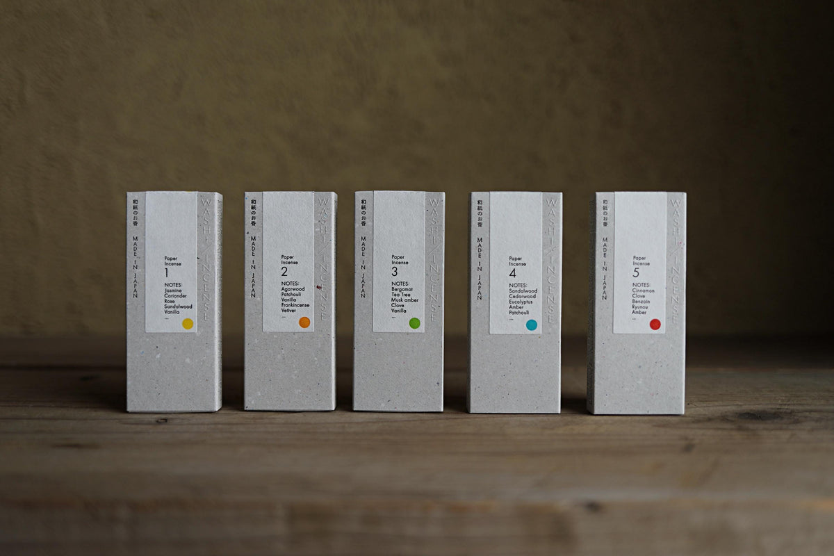 A row of five identical white boxes with colorful labels, each containing a different product name, arranged on a wooden surface.