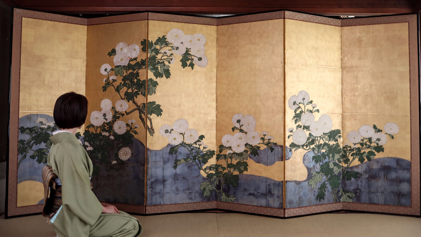 A person in a green kimono is kneeling in front of a large, intricately designed screen with floral and nature-inspired artwork.