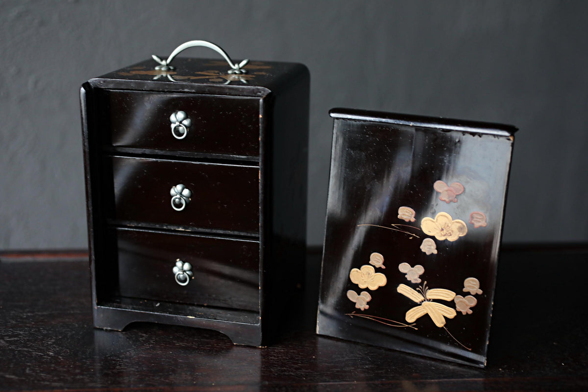 a black wooden box with three drawers and a handle, and a small black box with a floral design on it, both placed on a dark surface.