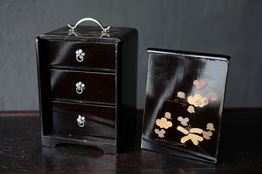 a black wooden box with three drawers and a handle, and a small black box with a floral design on it, both placed on a dark surface.