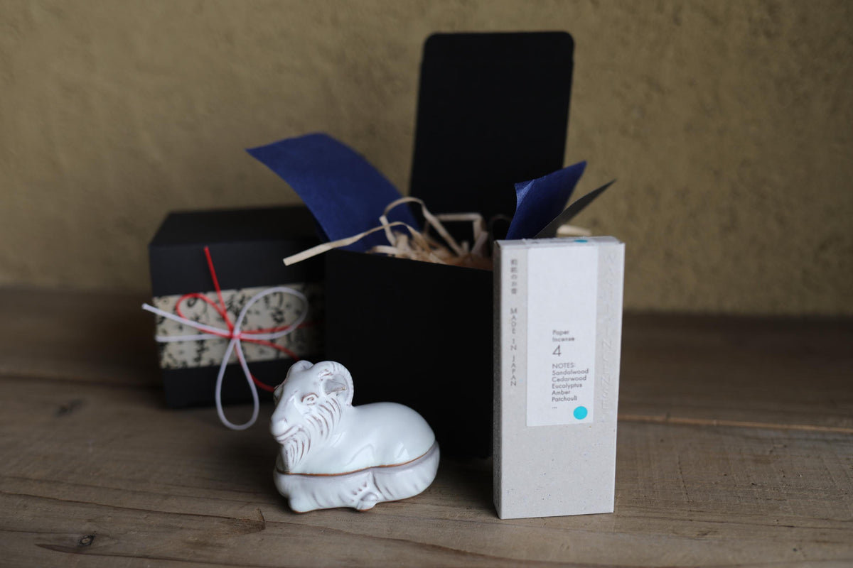 a white ceramic ram figurine, a black box, and a white box with blue and white text, all placed on a wooden surface.