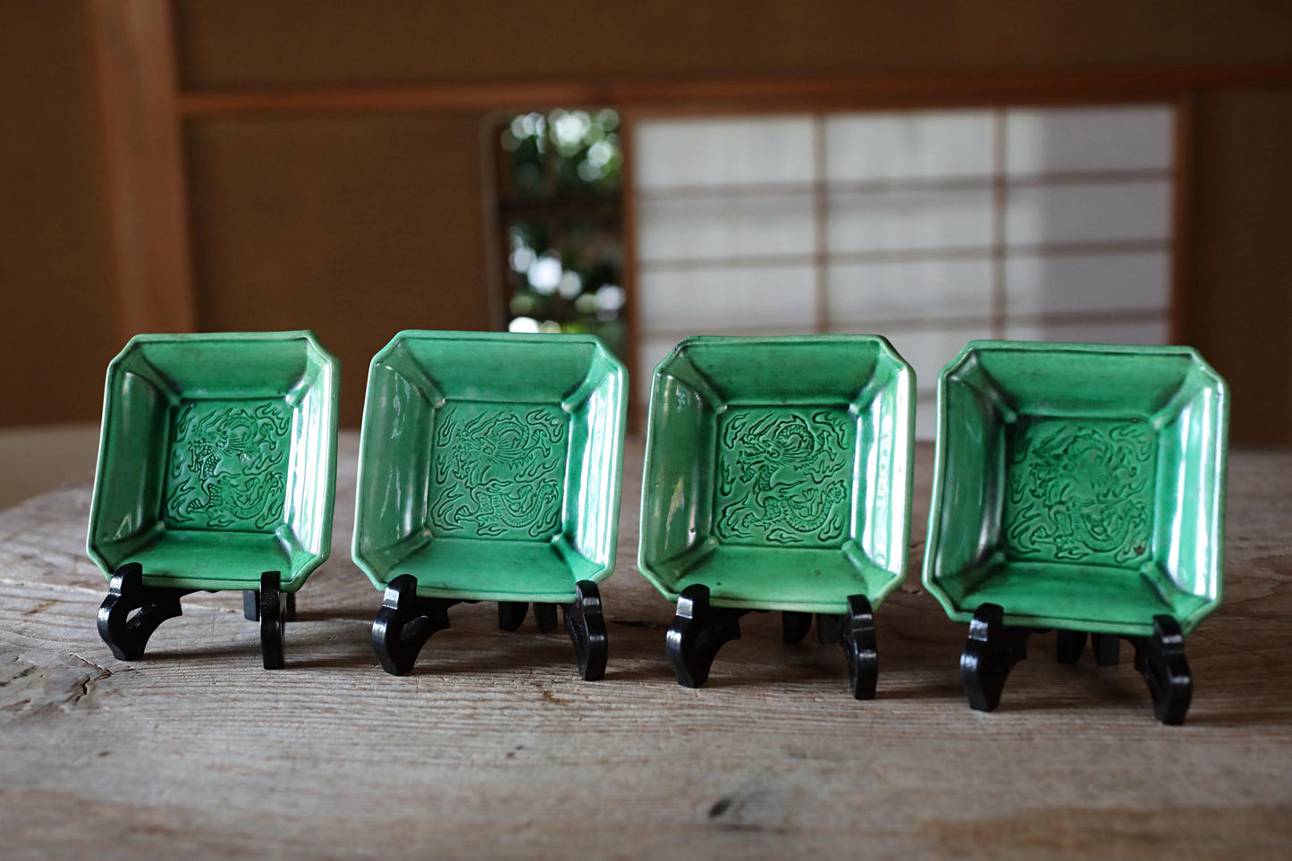 Four green ceramic plates with intricate designs are arranged on a wooden surface, each placed on a black stand.