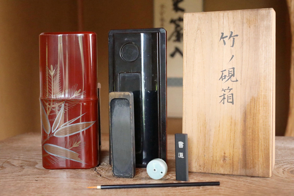 a collection of objects, including a red container with a floral design, a black container, and a wooden box with Japanese characters on it, all placed on a wooden surface.